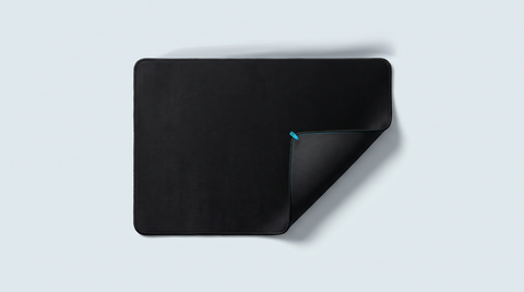 Grand tapis de bureau noir déroulé, un coin replié montrant sa surpiqûre et son dos caoutchouc